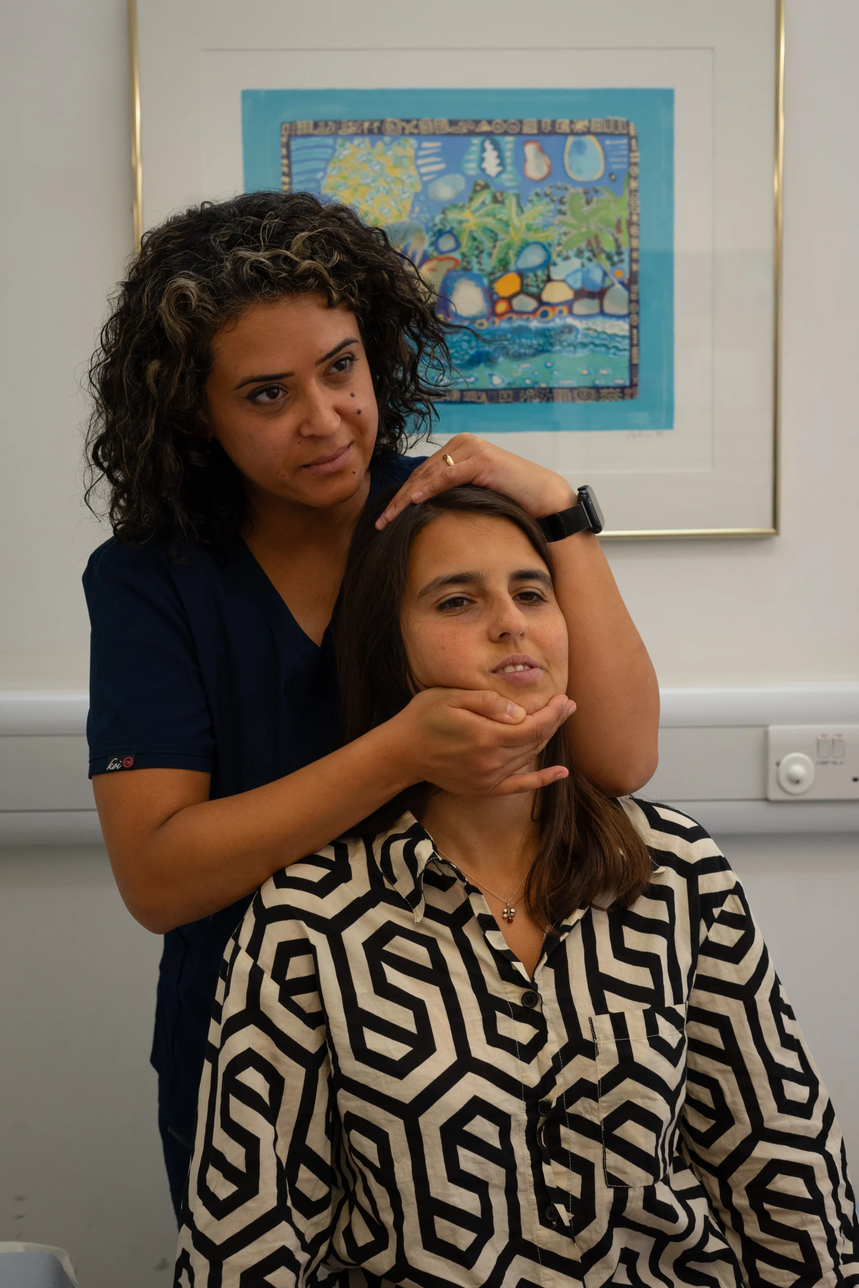 Caregiver stands behind a seated woman, gently holding her head and neck in a clinical room with a framed colorful artwork in the background.